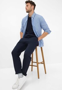 Lichtblauw geruit overhemd gedragen over een marineblauw T-shirt, gecombineerd met marineblauwe broek en witte sneakers, zittend op een houten kruk.