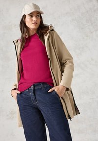 Beige wasserfeste Jacke mit Kapuze, über einem leuchtend pinken Pullover getragen. Kombiniert mit dunkelblauen, locker sitzenden Jeans. Hellfarbene Mütze.