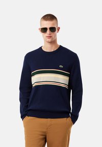 Suéter de punto azul marino con rayas horizontales en verde, crema, rojo y naranja, con cuello redondo y un logo de Lacoste en el pecho.