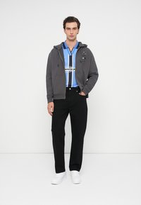 Γκρίζα φούτερ ζακέτα με λογότυπο και κορδόνια πάνω από μια μπλε και λευκή ριγέ πόλο μπλούζα, συνδυασμένη με μαύρο παντελόνι και λευκά αθλητικά παπούτσια.