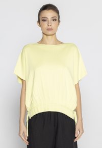 Żółta bluzka z krótkim rękawem z dzianiny, z prążkowanym dołem i sznurkami po bokach, w połączeniu z czarnymi, luźnymi spodniami.