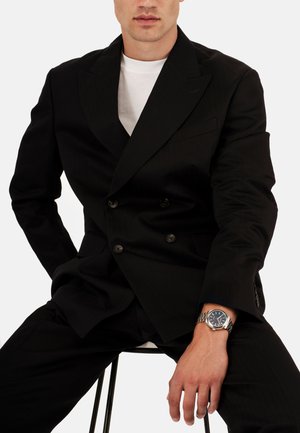 Veste de costume noire à double boutonnage avec des revers larges, associée à un t-shirt blanc et à un pantalon noir. Montre argentée au poignet, en pose assise.