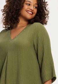 Olijfgroen tuniek top van een gestructureerde, gekreukte stof. Kenmerken een V-halslijn versierd met zilveren kraalaccenten en 3/4 mouwen met bell-sleeves.