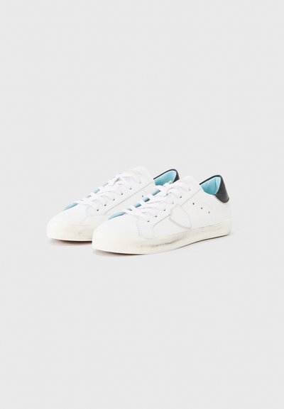 Baskets blanches avec accents bleus et noirs, dotées d'une tige en cuir lisse, d'un design à lacets et d'une semelle en caoutchouc usée.