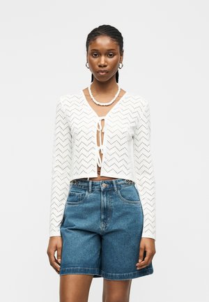 Femme portant un top blanc au crochet à manches longues avec des liens à l'avant, un short en jean bleu taille haute, un collier en coquillage et des boucles d'oreilles créoles.