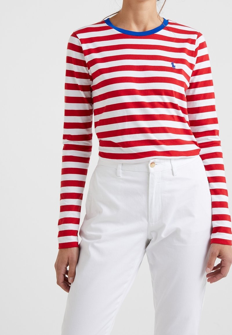 Maglietta a maniche lunghe a strisce rosse e bianche con colletto blu e piccolo logo sul petto, abbinata a pantaloni bianchi.