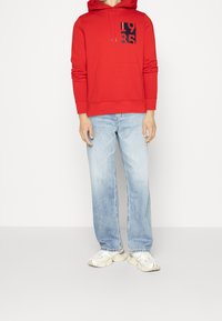 Punainen huppari, jossa on etupuolella kengurupussi, musta, rohkea graafinen "1985" -kuvio. Yhdistettynä vaaleansinisiin, väljäreunaisiin farkkuihin ja neutraaleihin lenkkareihin.