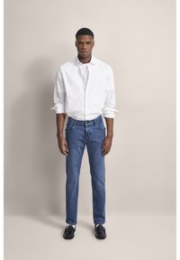 Biała koszula zapinana na guziki, niebieskie dżinsy oraz czarne buty. Slim-fit, gładka faktura i klasyczny niebieski kolor. Prosty styl.