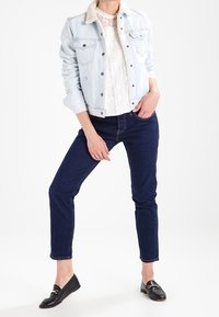 Giacca di jeans blu chiaro con collo in ecopelliccia, top in pizzo bianco, jeans blu scuro e mocassini neri con dettagli dorati.