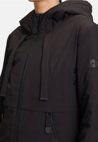 Černá zateplená bunda s kapucí, předn�ím zipem, nastavitelnými šňůrkami a knoflíkovými detaily. Materiál má hladkou matnou texturu.