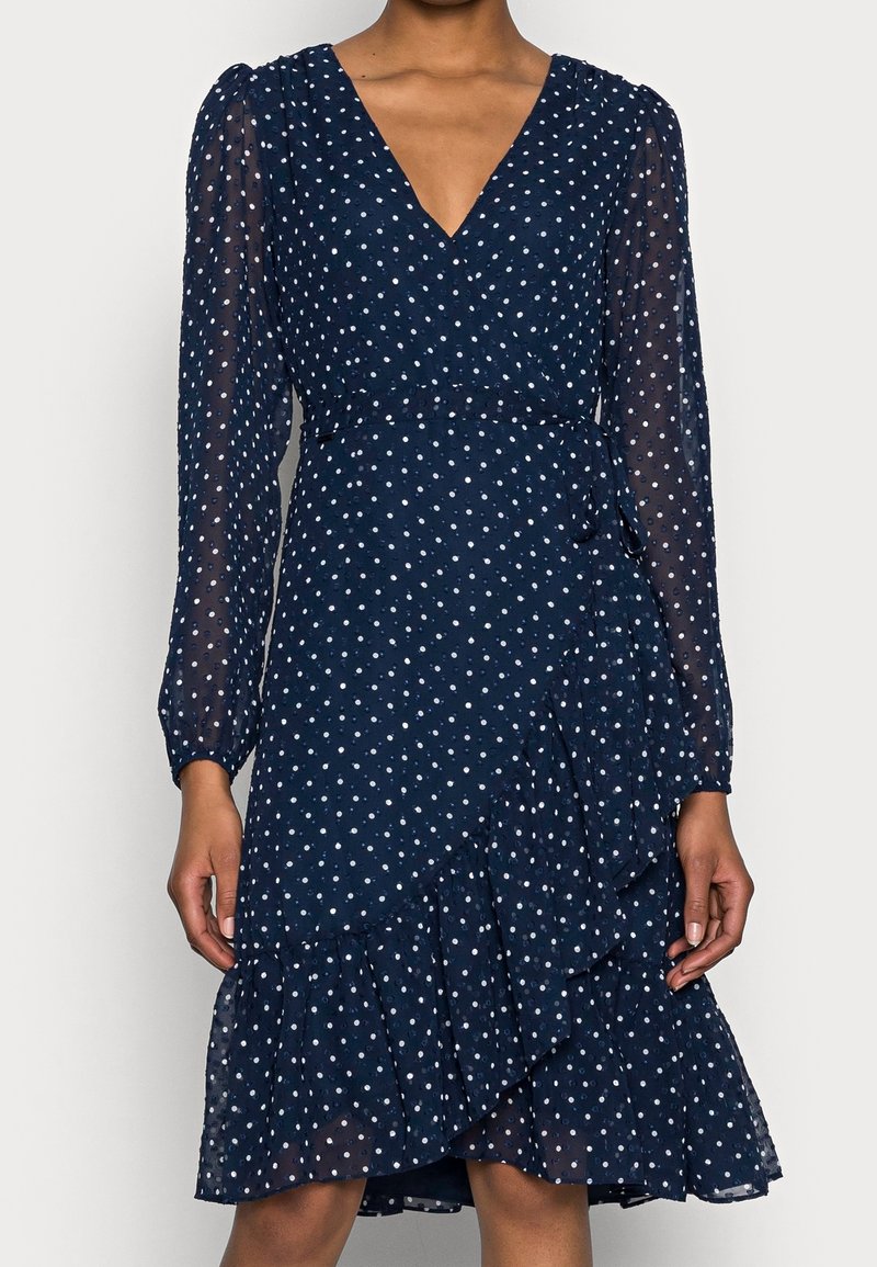 Tummansininen kietaisumekko, jossa on valkoisia pilkkuja, pitkät läpikuultavat hihat, V-kaula-aukko ja kerroksellinen röyhelöhelma.