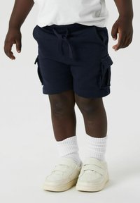 Shorts cargo bleu marine avec poches latérales, taille élastique et cordon de serrage. Associés à des chaussettes blanches côtelées et des baskets blanches à velcro.