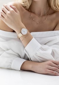 Montre en or rose avec un cadran blanc minimaliste et un bracelet à maillons, associée à des colliers délicats en or et gemmes, sur une blouse blanche.