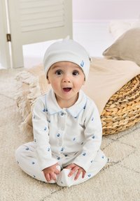 Witte katoenen peuter pyjamaset met blauwe zeilbootpatronen, knopen aan de voorkant en een bijpassende muts. Zachte textuur, ronde kraag en aansluitende boorden.