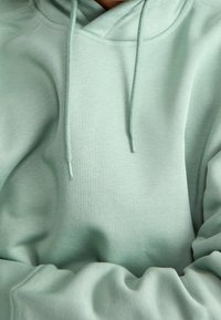 Mintgrön hoodie gjord av mjukt, texturerat tyg med dragsko i huvan och ribbade ärmar. Synliga sömmar förbättrar designen.