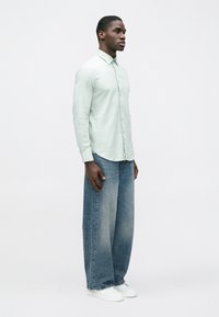 Šviesiai žalios spalvos ilgomis rankovėmis marškiniai su sagomis priekyje, derinami su laisvais, išblukusiais mėlynais džinsais ir baltais sportbačiais. Paprastas, kasdienis dizainas.