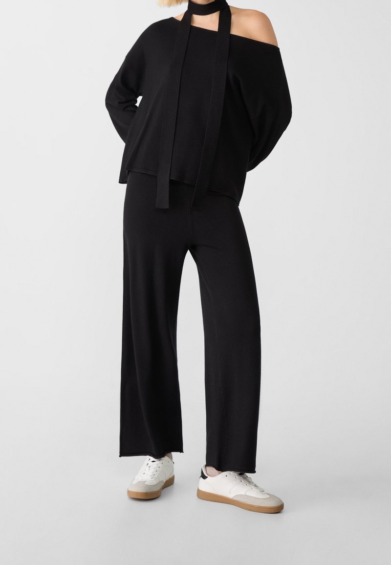 Personne portant un pantalon noir ample, un haut noir épaules dénudées avec de longs liens autour du cou, et des baskets blanches avec des semelles beige.