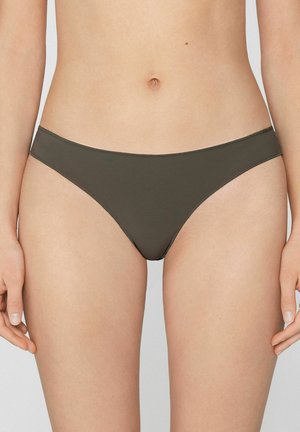 Kobieta nosząca proste, ciemnozielone majtki od bikini, pokazana od talii do połowy uda na neutralnym tle.