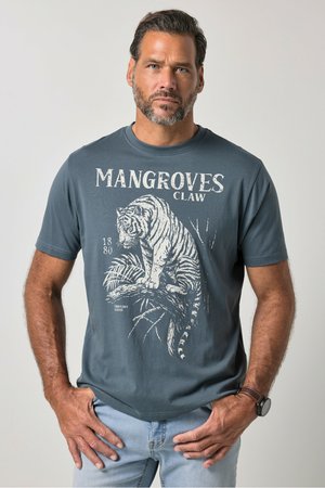 Mann trägt blau-graues T-Shirt mit weißem Tiger-Print und dem Text "MANGROVES CLAW", hellblaue Jeans und eine Armbanduhr.