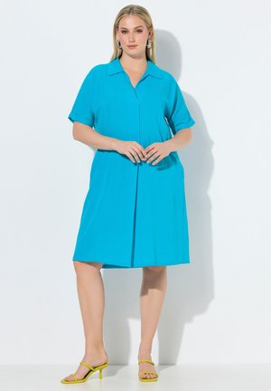 Kobieta o blond włosach ubrana w turkusową sukienkę do kolan z krótkim rękawem i żółte buty na obcasie z odkrytymi palcami, stoi na białym tle.