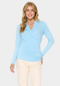 Šviesiai mėlynas ilgas rankovės marškinėliai su apvyniojimo dizainu ir V formos iškirpte. Minkšta medžiaga su glotnia tekstūra. Modelis dėvi šviesiai smėlio spalvos kelnes.