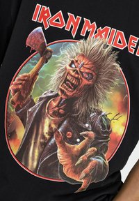 Camiseta negra con un gráfico de un espectro con cabello desordenado, sosteniendo un hacha. Fondo rojo y naranja con el texto "IRON MAIDEN" en letras grandes.