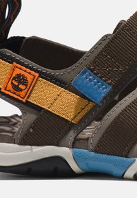 Sandalias deportivas marrones y grises con correas multicolor: acentos en naranja, amarillo y azul; suela texturizada y plantilla acolchada.