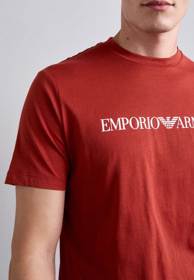 Vyras, dėvintis raudoną trumpų rankovių marškinėlį su baltu „Emporio Armani“ logotipu, atspausdintu ant krūtinės, kirptu kaklo ir vidurinėje rankovėje.
