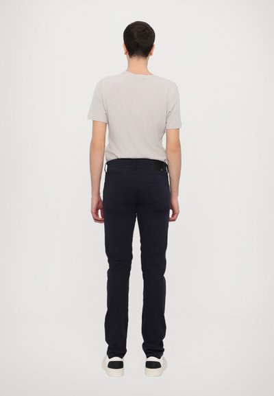 Γκρι κοντομάνικο μπλουζάκι συνδυασμένο με παντελόνι ναυτικού χρώματος, με κομψή εφαρμογή και ελάχιστες λεπτομέρειες. Το μοντέλο στέκεται στραμμένο προς τα πίσω, φορώντας λευκά αθλητικά παπούτσια.