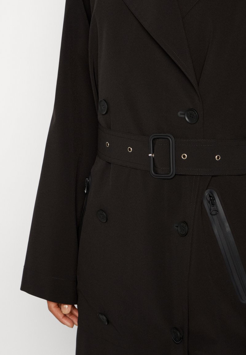 Cappotto nero doppiopetto con vita cinturata, grandi bottoni decorativi e tasche laterali con zip. Tessuto testurizzato con una finitura liscia.