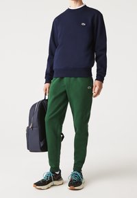 Felpa navy, pantaloni sportivi verdi, scarpe nere con accenti colorati e uno zaino navy. Presenta loghi sul petto e sui pantaloni.