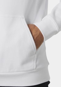 Mano de una persona con sudadera blanca descansando dentro del bolsillo frontal tipo canguro sobre un fondo liso.