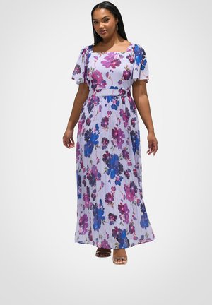 Kvinde iført en hvid maxikjole med blomsterprint i pink, blå og lilla, flagrende ærmer og firkantet udskæring, stående mod en ensfarvet baggrund.