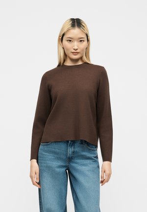 Jeune femme aux cheveux blonds et raides portant un pull marron à manches longues et un jean bleu, debout devant un fond blanc uni.