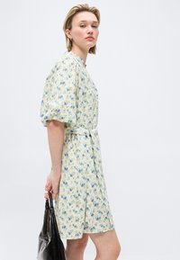 Γυναίκα με ανοιχτό κίτρινο φόρεμα με λουλούδια, φουσκωτά μανίκια και κορδέλα στη μέση, κρατώντας μια μαύρη τσάντα, σε απλό λευκό φόντο.