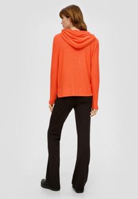 Oranje hoodie met een ontspannen pasvorm, zachte textuur en lange mouwen, gecombineerd met zwarte flare-broeken en dikke zwarte schoenen.