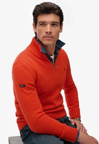 Maglione lavorato a maglia arancione con colletto alto, polsini a costine e chiusura con zip. Indossato sopra una camicia a quadri blu, con un logo sulla manica.