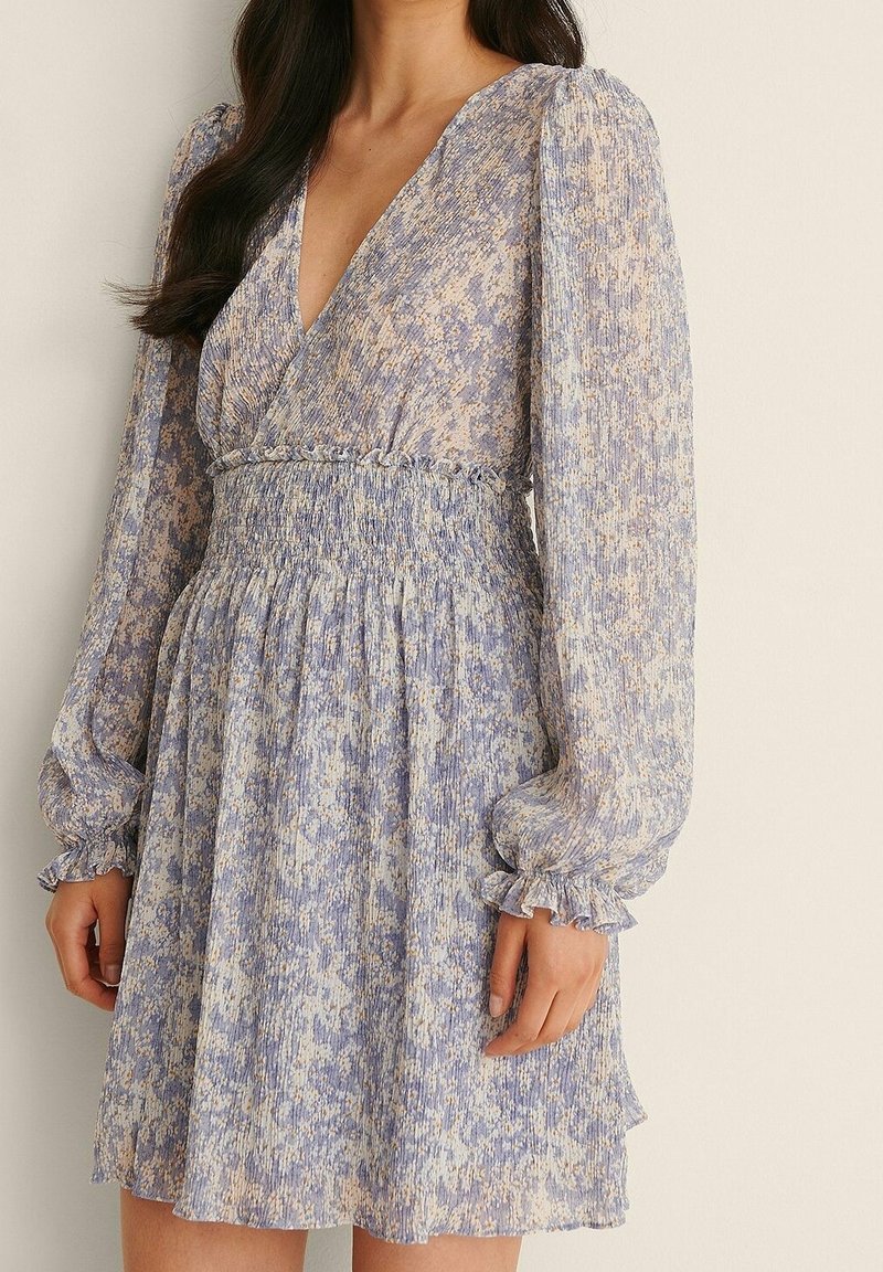 Femme portant une robe à manches longues, col en V, imprimé floral, avec une taille smockée et des poignets volantés, debout devant un fond uni.