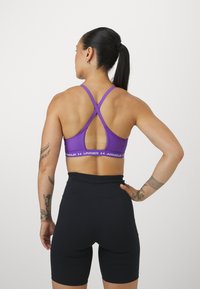 Sostenedor deportivo púrpura con tirantes entrecruzados y banda con logo, combinado con pantalones cortos negros de cintura alta. Tejido suave, ajuste ceñido al cuerpo.