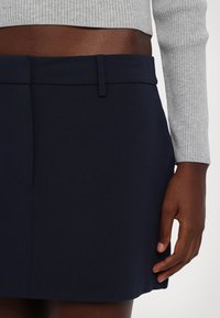 Jupe bleu marine en tissu lisse, avec une ceinture plate, des poches latérales et une coupe droite. Gros plan sur une main posée sur le tissu.