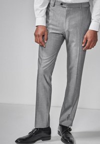 Hellgraue Anzughose aus glattem Stoff mit tailliertem Schnitt, im Flat-Front-Design, seitlichen Taschen und Gürtelschlaufen. Getragen mit schwarzen Schuhen.