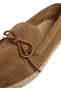 Mocassin en daim marron avec un bout rond, des détails de lacets en cuir et une semelle en jute tissée. Présente un emblème de marque sur le côté.