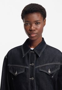 Tumši zils džinsa žakete ar smailu apkakli, divām krūšu kabatām, sudraba detaļām un baltu izšuvumu, radot strukturētu dizainu.