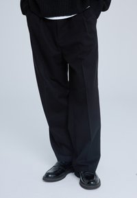 Pantaloni neri su misura con design a gamba dritta e pieghe. Realizzati in un tessuto testurizzato, abbinati a mocassini neri lucidi.