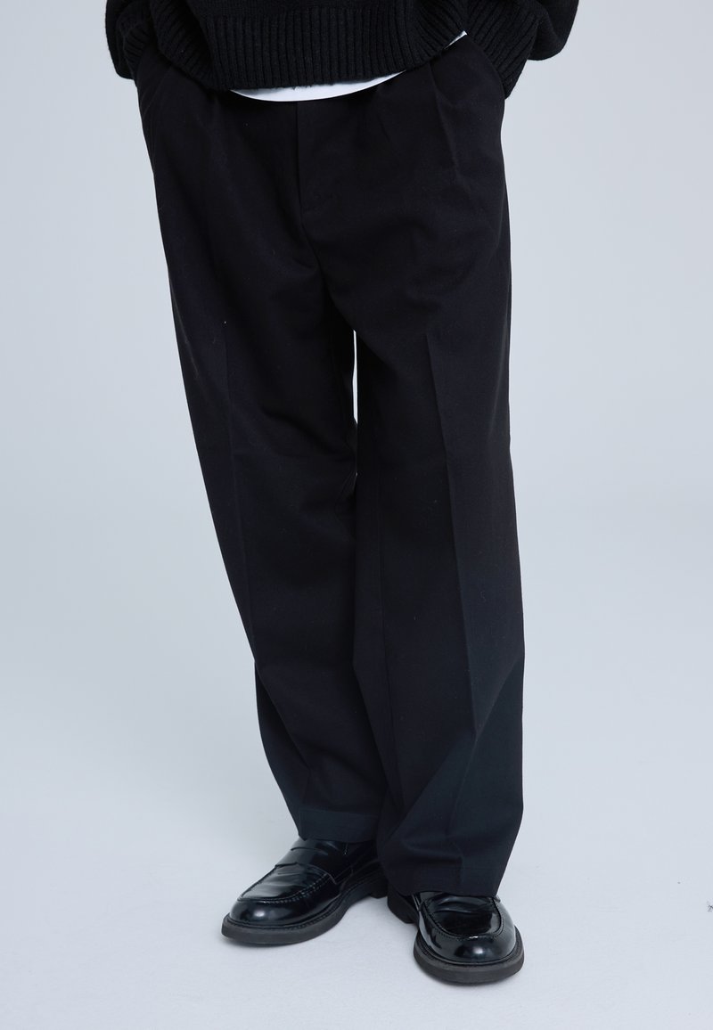 Pantaloni neri su misura con design a gamba dritta e pieghe. Realizzati in un tessuto testurizzato, abbinati a mocassini neri lucidi.