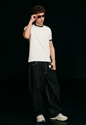 Mladý muž si upravuje slnečné okuliare, má na sebe biele tričko s farebným lemovaním, voľné čierne džínsy s príveskom na kľúče a biele tenisky, stojí pred čiernym pozadím.