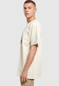 Cremefarvet bomulds t-shirt med korte ærmer, løs pasform, med sort broderet tekst på forsiden. Glat tekstur, minimalistisk design.