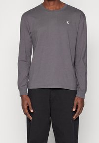 Harmaa pitkähihaista t-paitaa, joka on valmistettu puuvillasta ja jossa on miehusta sekä pieni logo. Hiukan istuva malli, jossa näkyvät saumat.