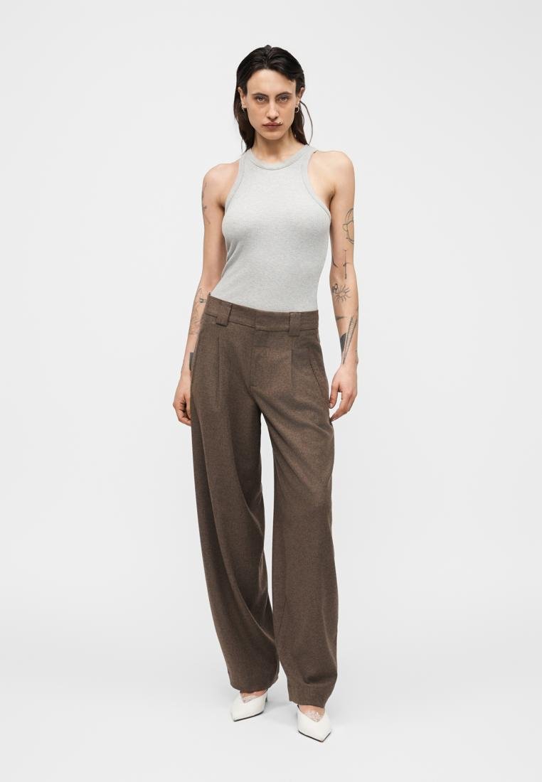 Femme aux cheveux foncés et tatouages, vêtue d'un haut gris sans manches, d'un pantalon large marron et de talons blancs pointus, debout devant un fond uni.