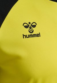 Tissu de sport jaune et noir avec un logo stylisé d’abeille noire au-dessus du nom de la marque « hummel » imprimé dessus.